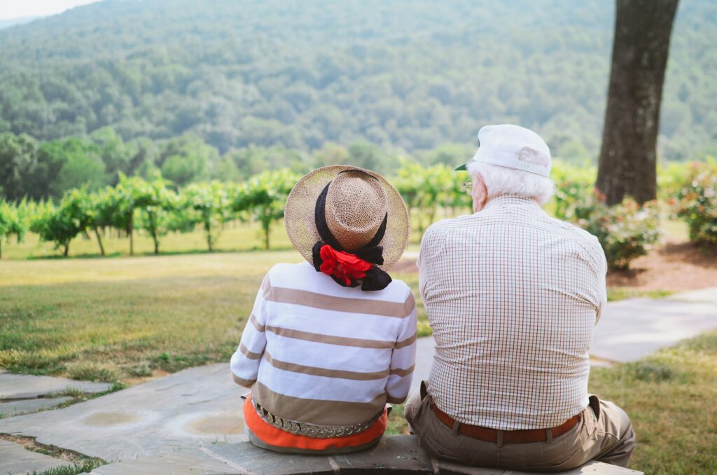 An elderly couple sitting outdoors on a sunny day, enjoying a peaceful view of a vineyard and lush green hills, symbolizing the benefits of staying at home with personalized care.