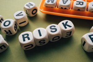 The image shows several wooden blocks with letters on them, spelling out the word "RISK" in a prominent row. Other blocks are scattered around, some partially showing letters. There's an orange tray holding some of the blocks, and the background is a plain light green surface. The image conveys themes of risk or uncertainty, often associated with decision-making or evaluation.